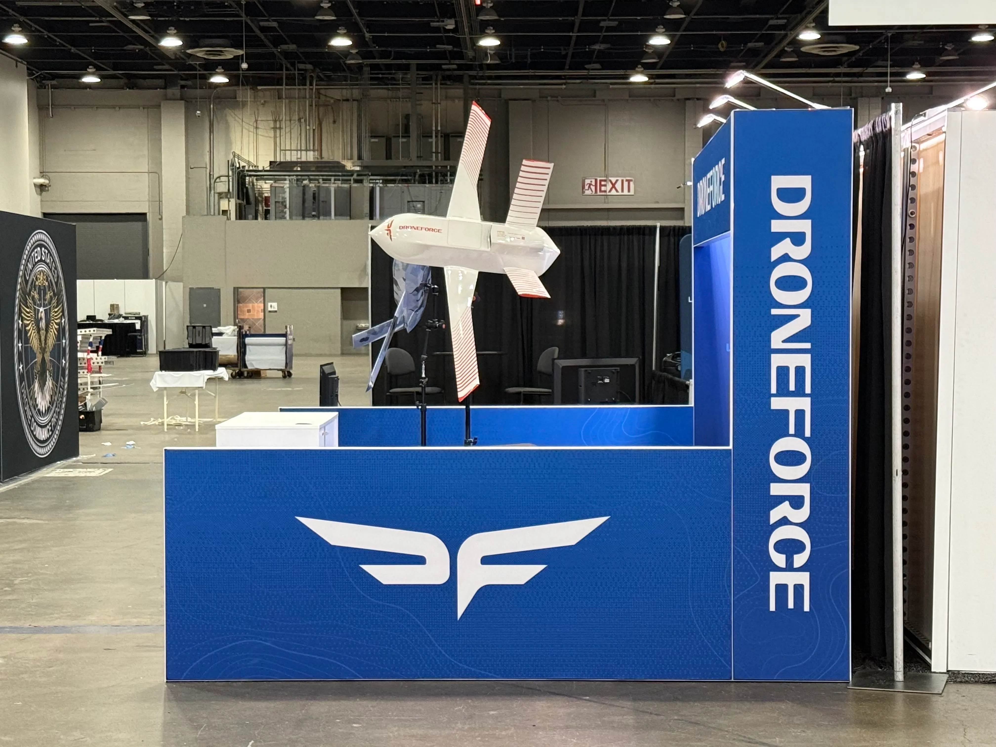 A white quadplane drone with red-striped propellers on the Droneforce booth display stand.