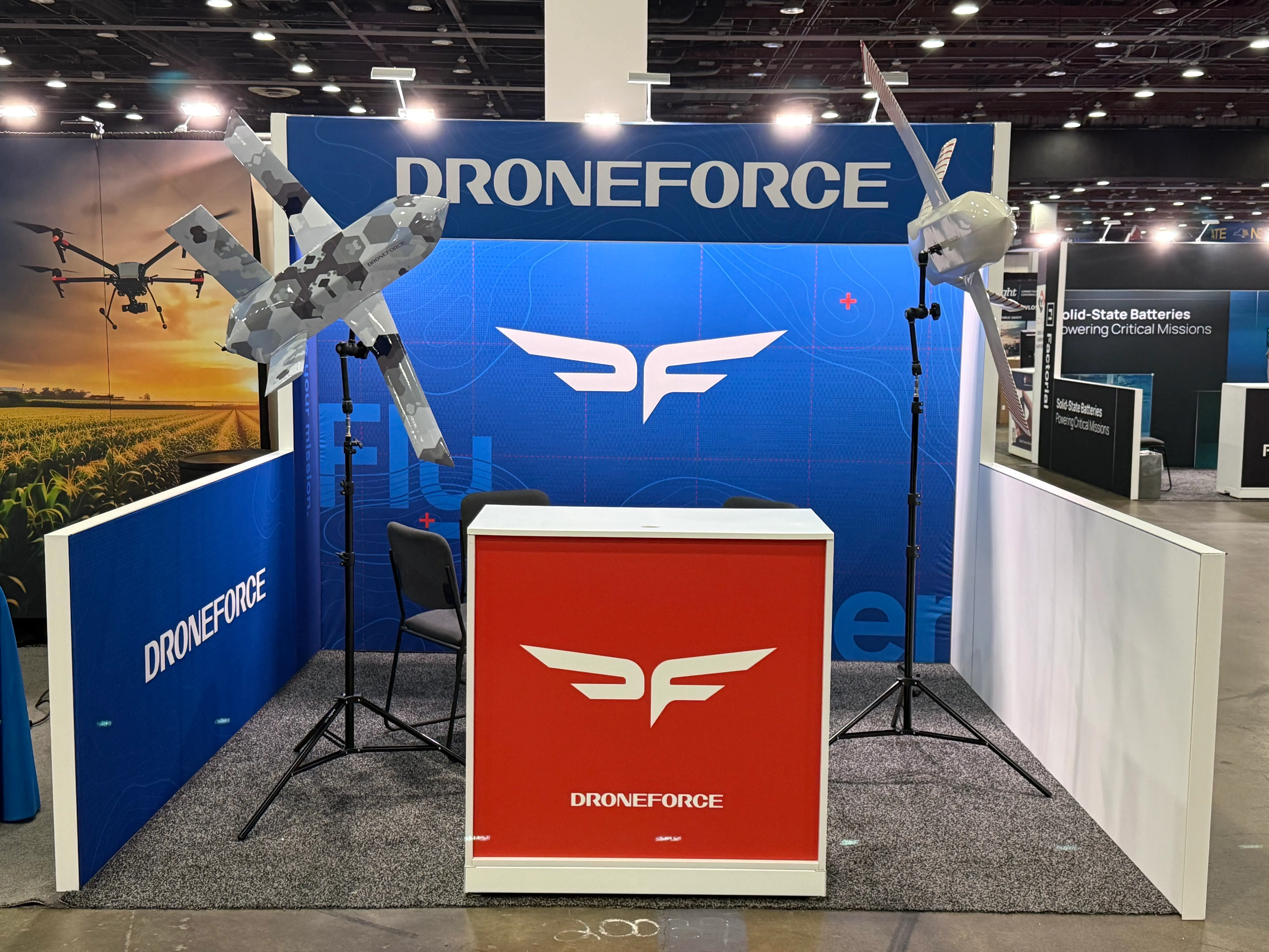 Two Droneforce fixed-wing aircraft on display stands at the booth, one in hexagonal camouflage, one in white.